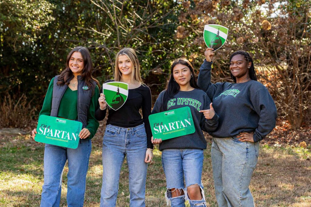 group photo of 4 students holding USC Upstate signs with the green trees of campus in the background